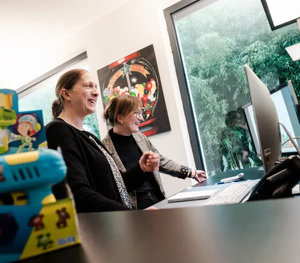 Zwei Frauen lächeln und schauen auf einen Computerbildschirm, während eine farbige Spielzeugfigur im Vordergrund zu sehen ist. Im Hintergrund hängt ein Bild mit Bonbons.