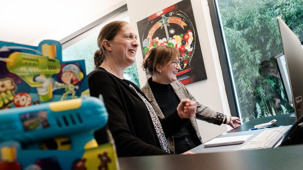 Zwei Frauen lächeln und sehen auf einen Computerbildschirm, im Vordergrund ist ein buntes Spielzeug sichtbar. Im Hintergrund ist eine grüne Pflanzenlandschaft zu erkennen.