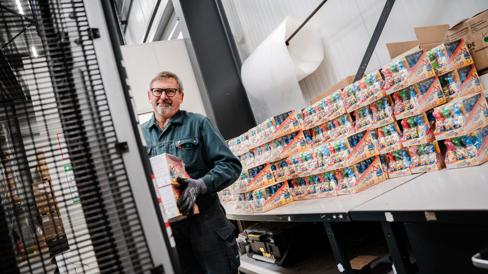 Ein Mitarbeiter in einem Lager hält eine Kiste mit Produkten, während im Hintergrund stapelweise Verpackungen zu sehen sind.