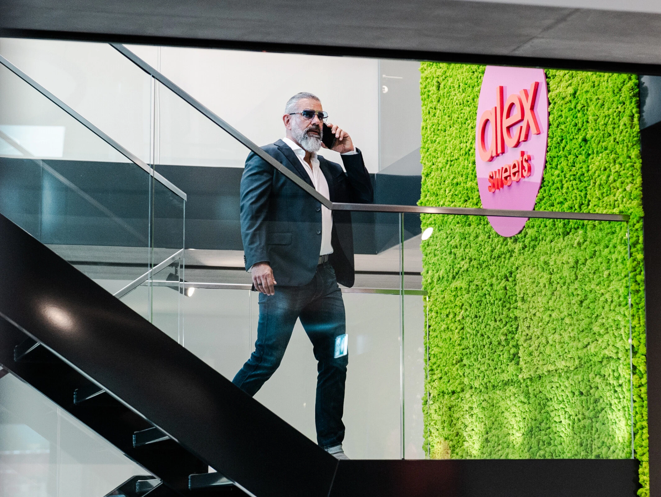 Ein Mann in einem Anzug mit Sonnenbrille spricht am Telefon und geht eine Treppe in einem modernen Raum mit einer grünen Wand und dem Schriftzug „alex sweets“ hinunter.