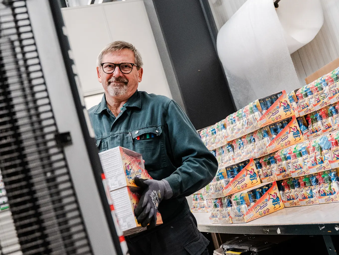 Mann mit Arbeitshandschuhen und einer Brille lächelt, während er Kartons mit Spielzeug verräumt