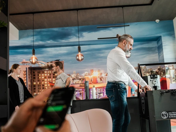 Büro mit mehreren Personen, die miteinander kommunizieren und ein Tischfussballspiel aufstellen. Modernes Ambiente mit Stadtlandschaft im Hintergrund.