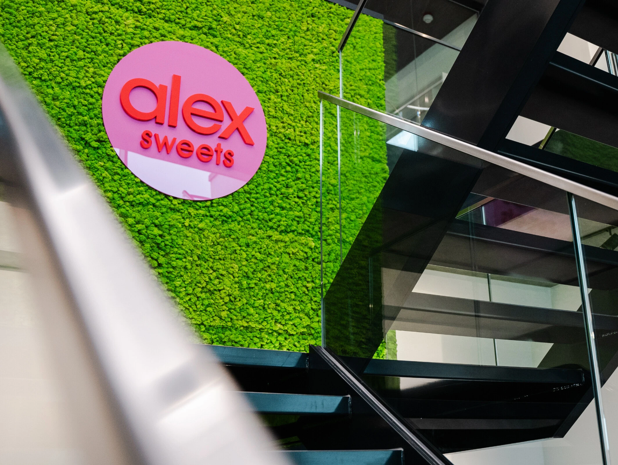 Moderne Treppe mit Glasgeländer und einer hellgrünen Mooswand. Das Logo „alex sweets“ in rosa ist sichtbar.