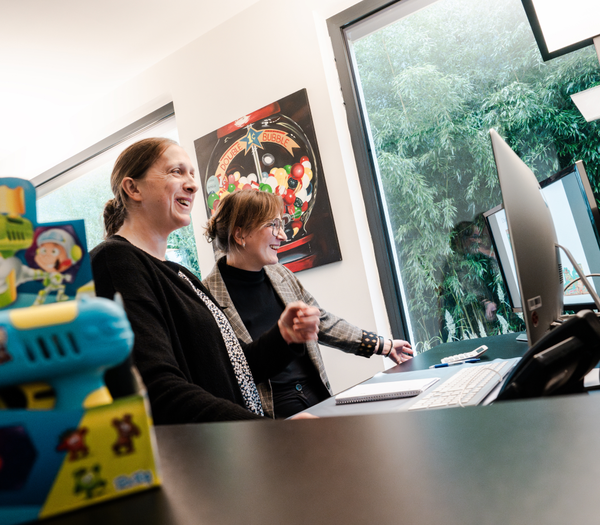 Two women are smiling and looking at a computer screen, while a coloured toy figure can be seen in the foreground. A picture of sweets hangs in the background.