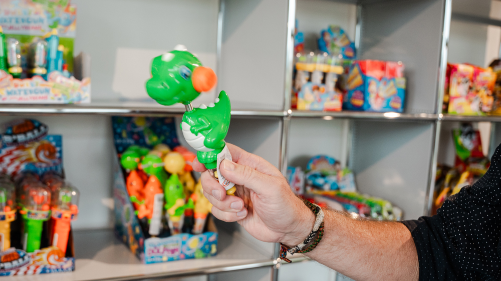 A green dinosaur toy with an orange ball, held by a person, in front of a shelf full of colourful toys.