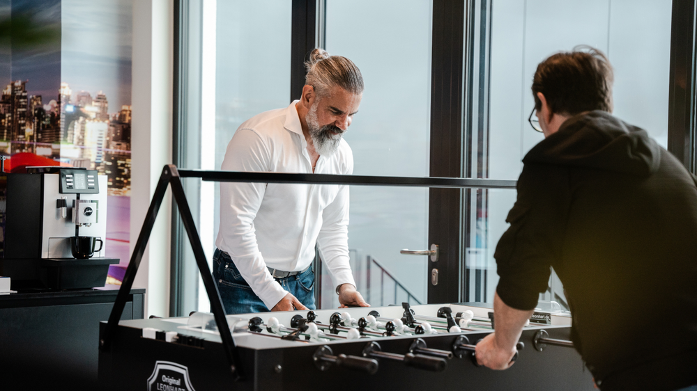 Two men play table football in a modern room with a window view. A coffee machine stands next to it.