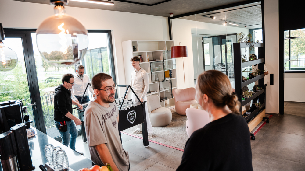 A modern office or living environment with several people chatting. The focus is on a table football game and cosy seating. Large windows offer a view of nature.