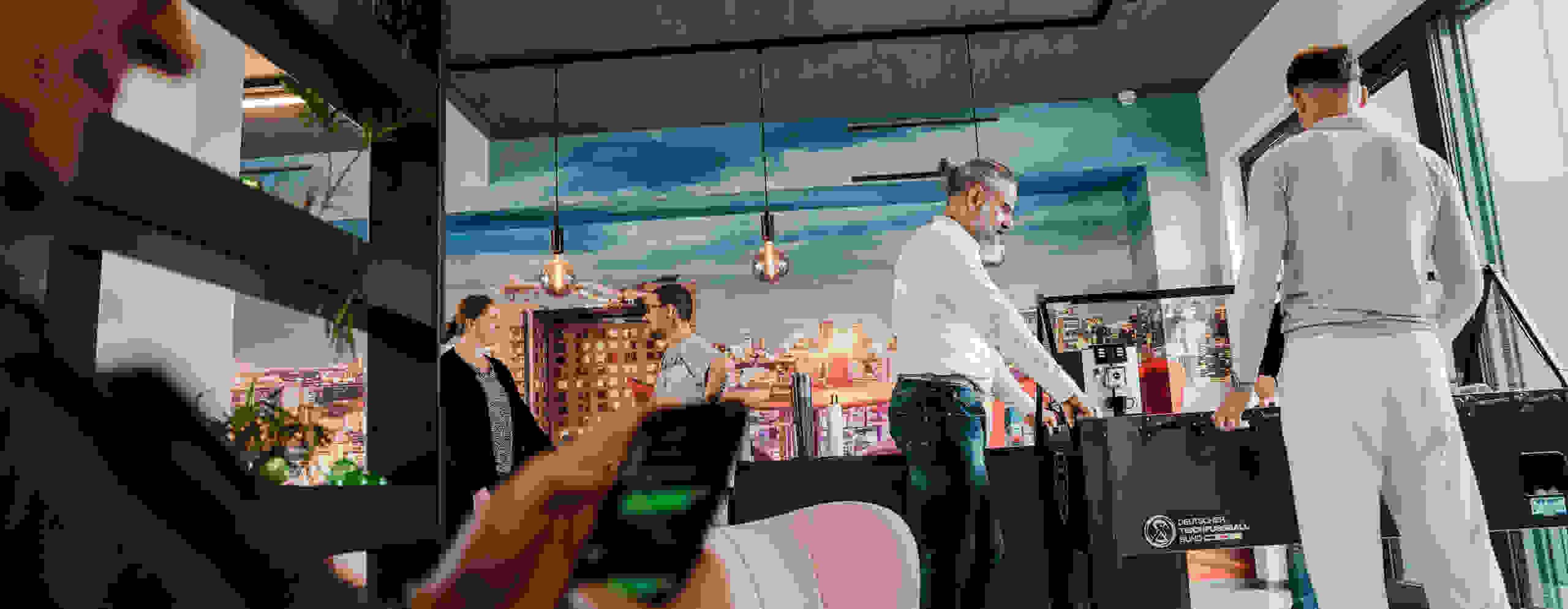An employee playing table football in a modern office with cityscape decor, while other colleagues are talking and another person is typing on a smartphone.