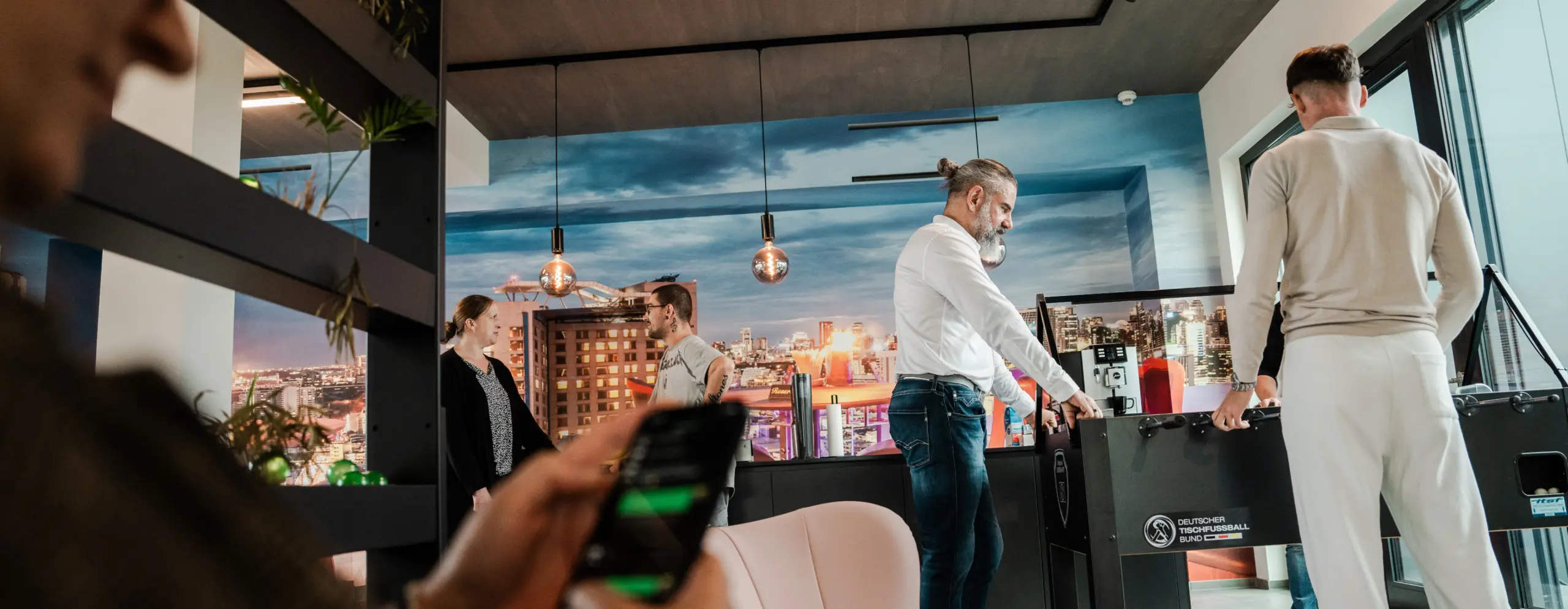 An employee playing table football in a modern office with cityscape decor, while other colleagues are talking and another person is typing on a smartphone.