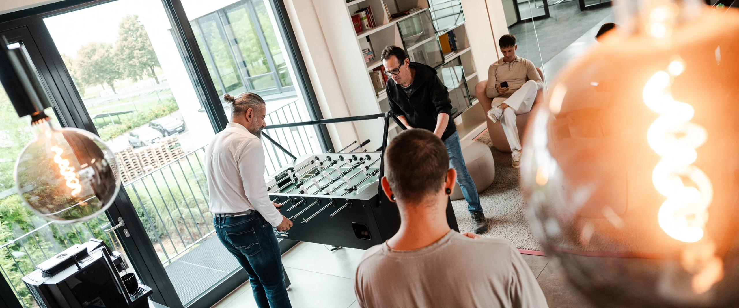 Employees playing table football in a modern, bright office lounge, while another colleague sits on a chair using their phone.