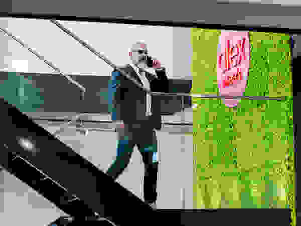 A man in a suit with sunglasses talks on the phone and walks down a staircase in a modern room with a green wall and the words 