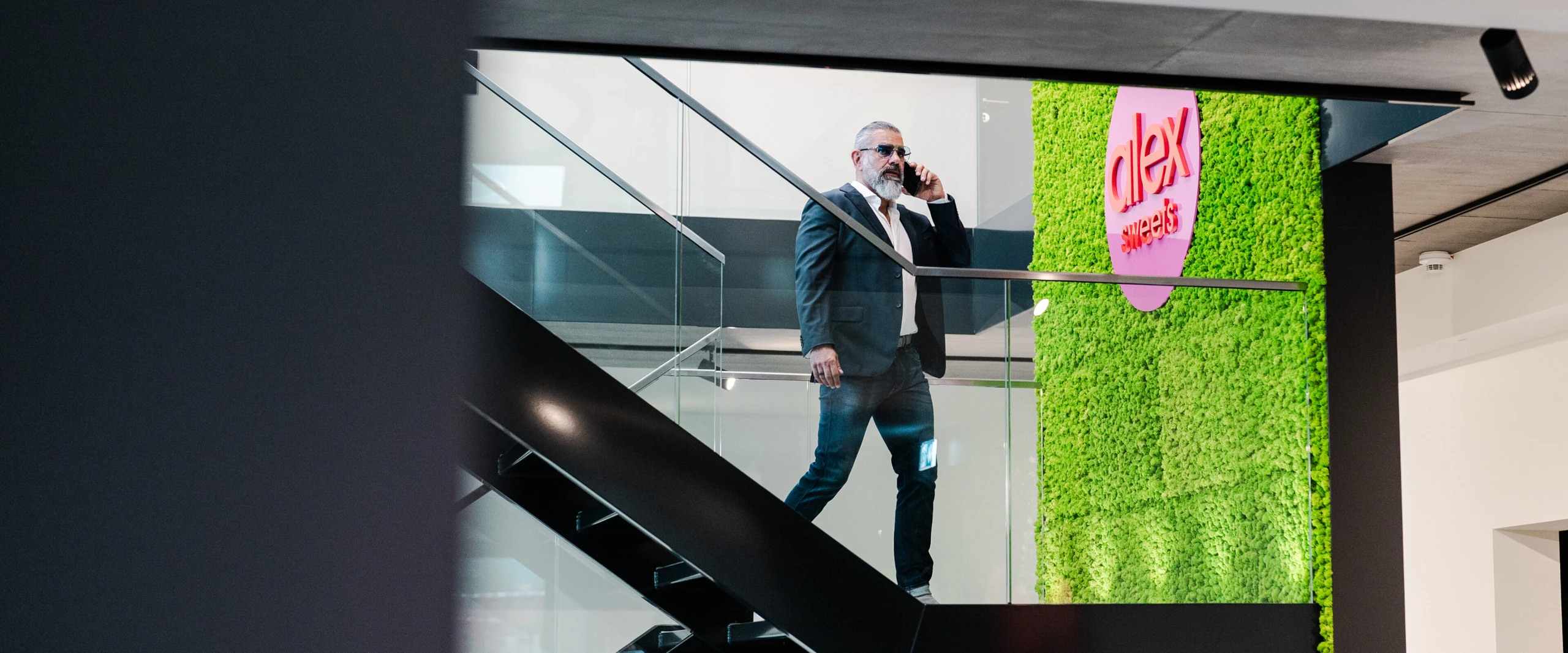 Un hombre trajeado con gafas de sol habla por teléfono y baja una escalera en una habitación moderna con una pared verde y las palabras 
