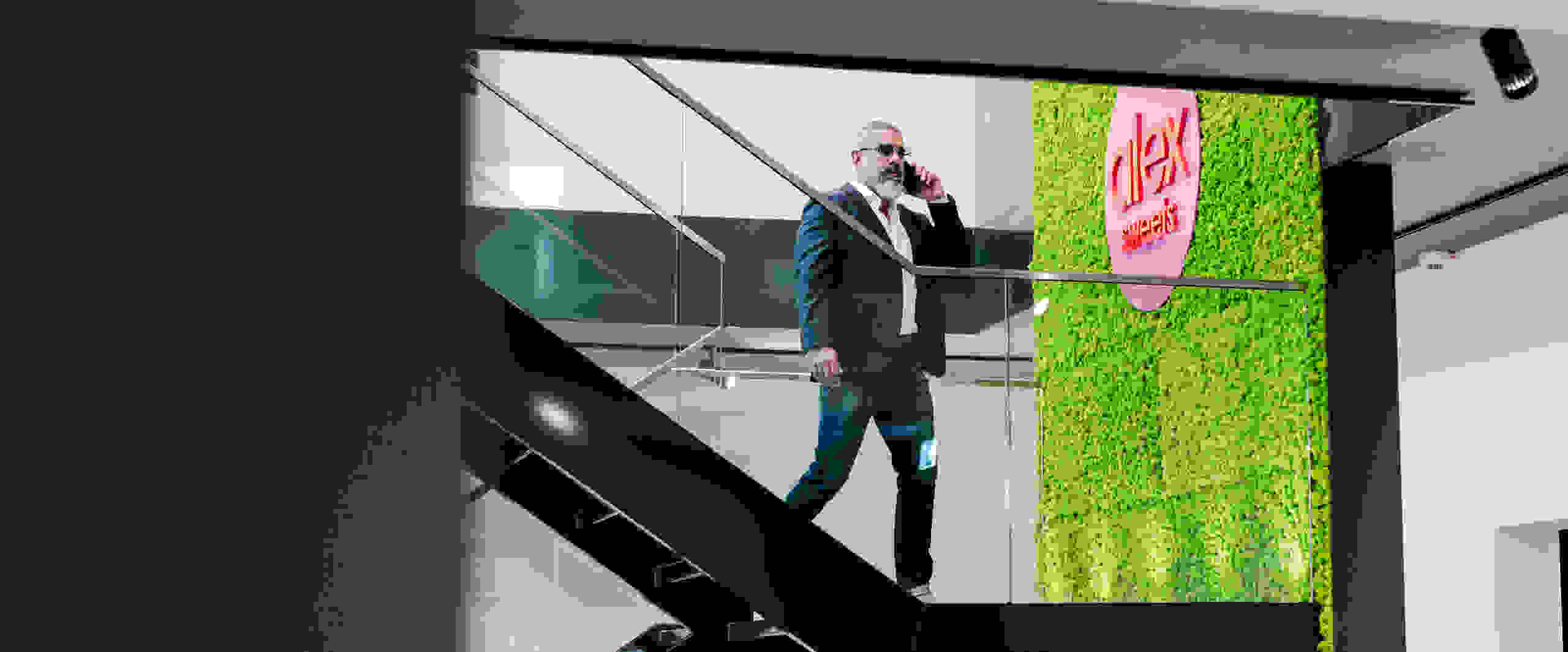 Un hombre trajeado con gafas de sol habla por teléfono y baja una escalera en una habitación moderna con una pared verde y las palabras 