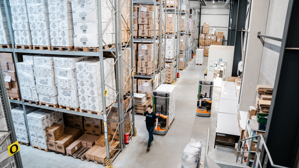 Un entrepôt avec de hautes étagères remplies de cartons anguleux. Un employé se déplace dans l'allée, tandis qu'un véhicule robot est visible en arrière-plan.