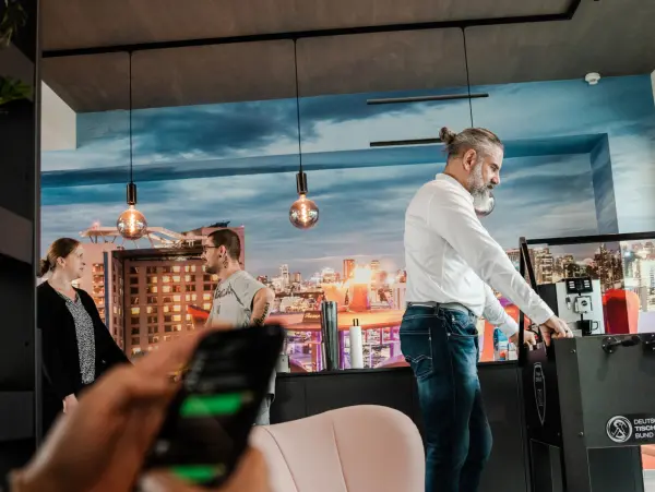 Un employé joue au baby-foot dans un bureau moderne avec une décoration de paysage urbain, tandis que d'autres collègues discutent et qu'une autre personne tape sur un smartphone.