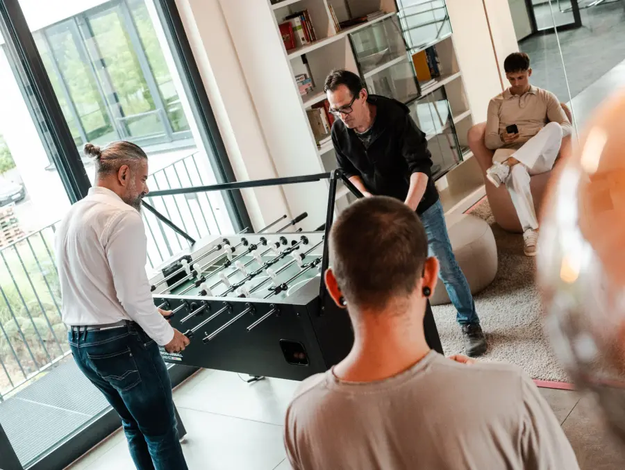 Dipendenti che giocano a calcio balilla in una lounge d'ufficio moderna e luminosa, mentre un altro collega è seduto su una poltrona e utilizza il suo telefono.